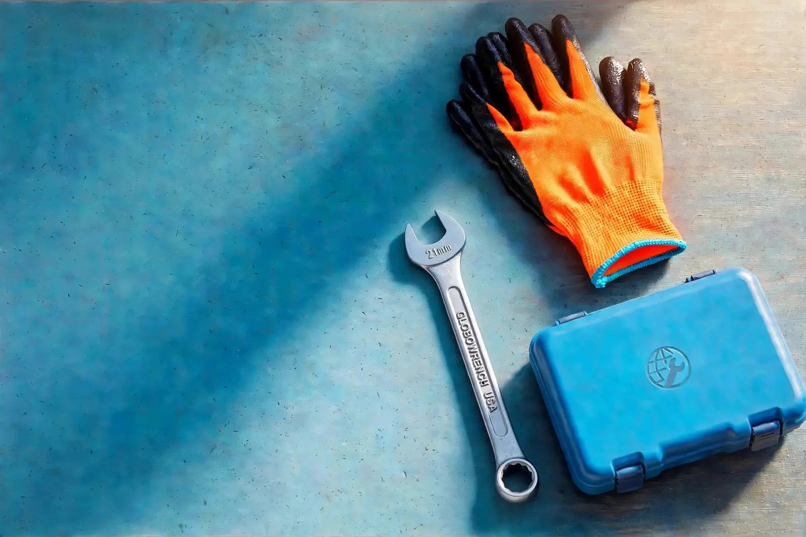 Thin hi-viz orange work gloves, a large all-metal 21mm combination wrench stamped GLOBOWRENCH USA, and a cyan tool case on a richly textured blue workshop surface